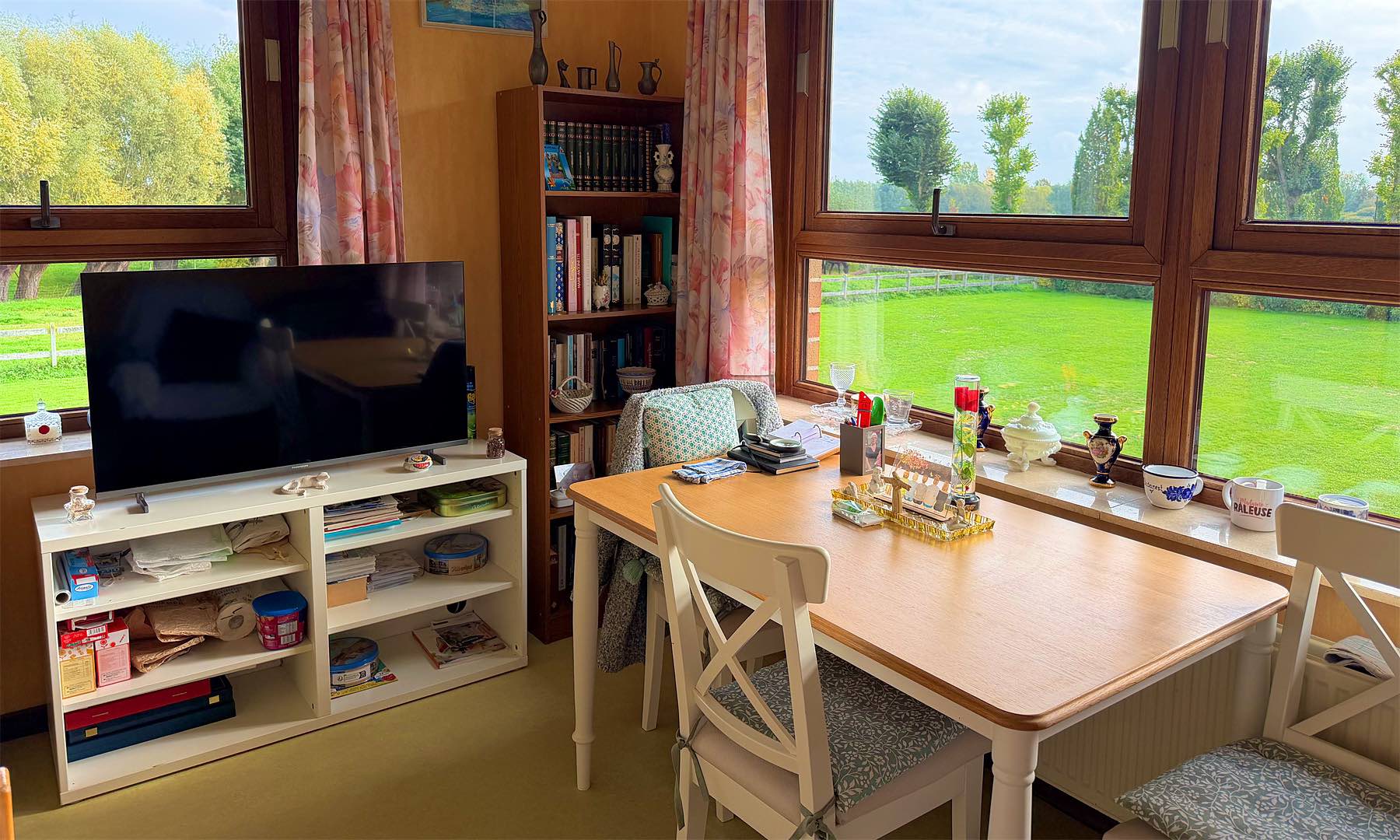 Chambre individuelle : table, télévision, bibliothèque et grandes fenêtres sur la campagne