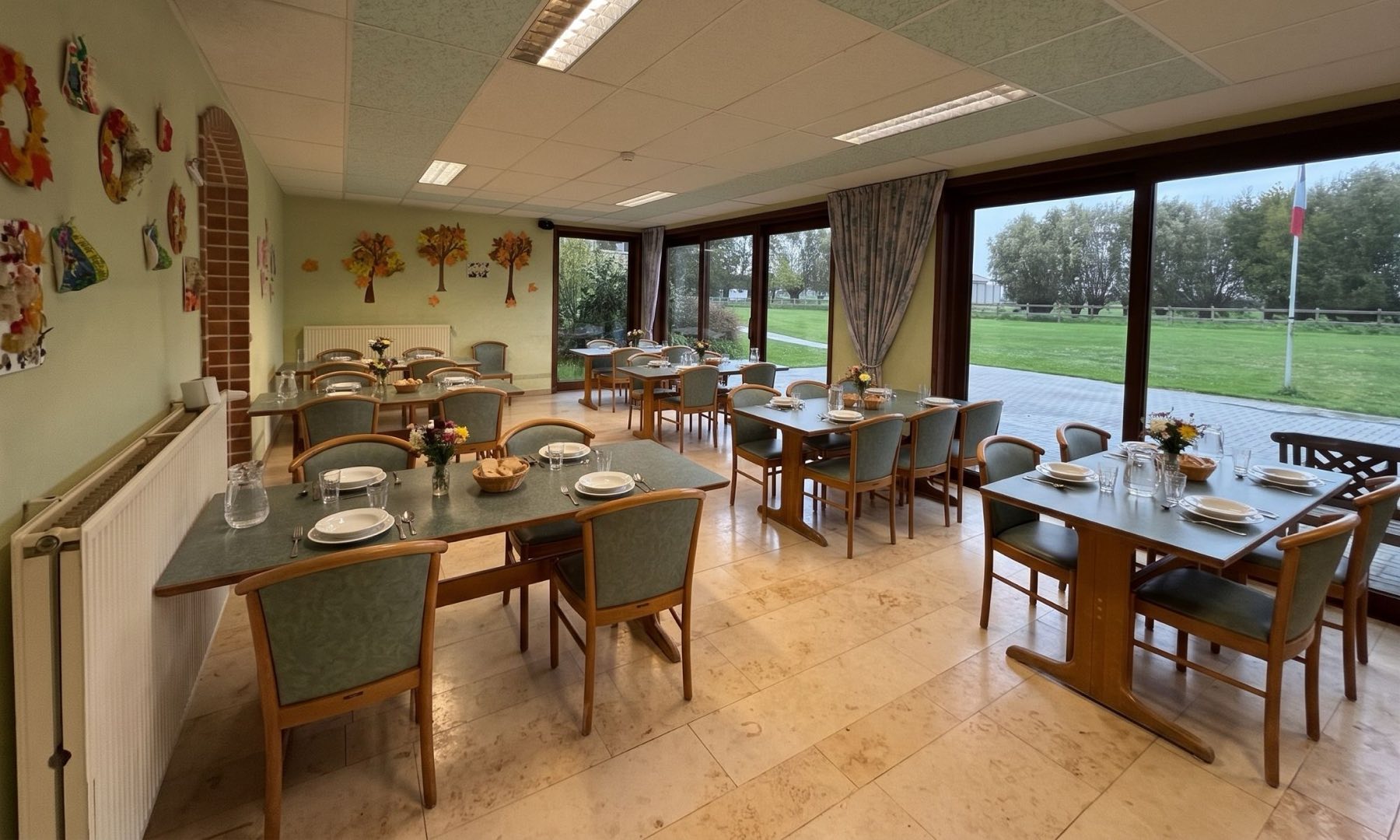 La salle à manger, ses tables dressées et ses grandes baies vitrées ouvertes sur la pelouse